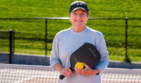 A woman smiling while holding a pickleball and paddle.