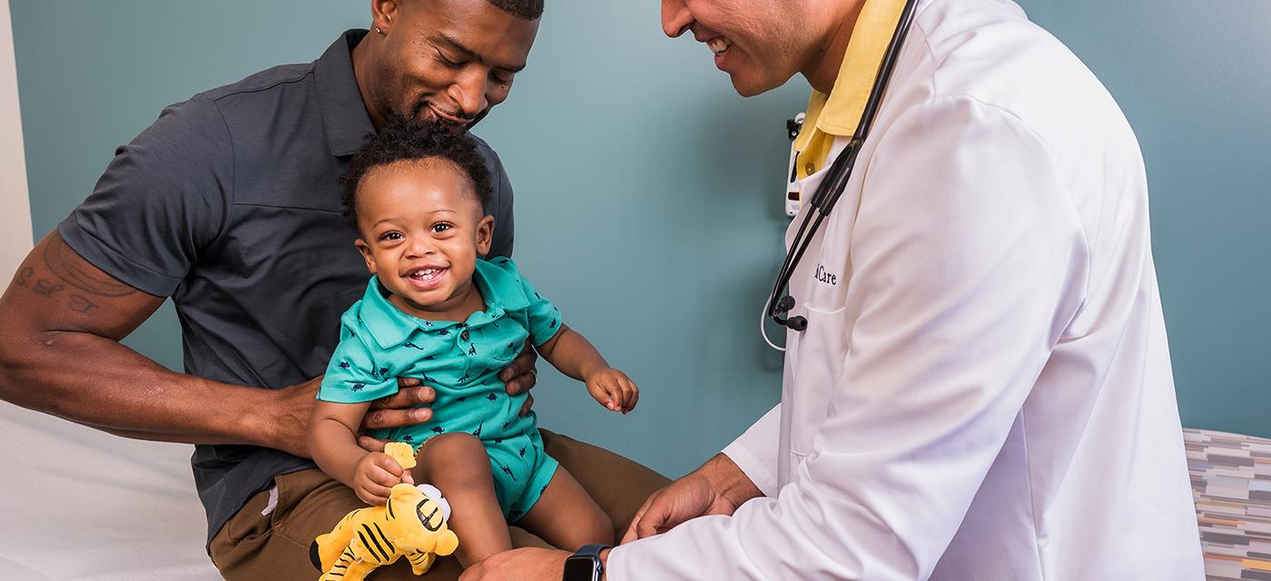 A father holding his son, while a primary care doctor checks the child's vitals.